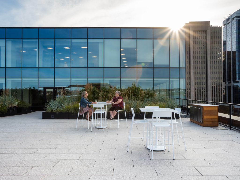 Two women chat while sitting on white Foro Bar height stools at a table in front of a large planter box